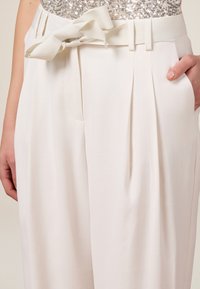 Pantalon blanc sur mesure à jambe large, doté d'un accent de nœud sur la taille et de plis à l'avant, fabriqué dans un tissu lisse.