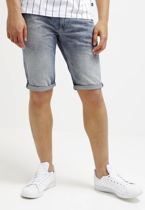Personne portant un short en denim léger retroussé, des baskets blanches et une chemise blanche à rayures verticales noires, debout sur un fond blanc.