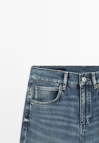 Sezione vita di jeans di denim blu con tasca frontale, passanti per cintura e chiusura con bottone metallico su sfondo bianco.