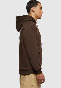 Sudadera de algodón marrón con un bolsillo canguro, capucha con cordón y puños de canalé, mostrada en un modelo masculino de perfil.