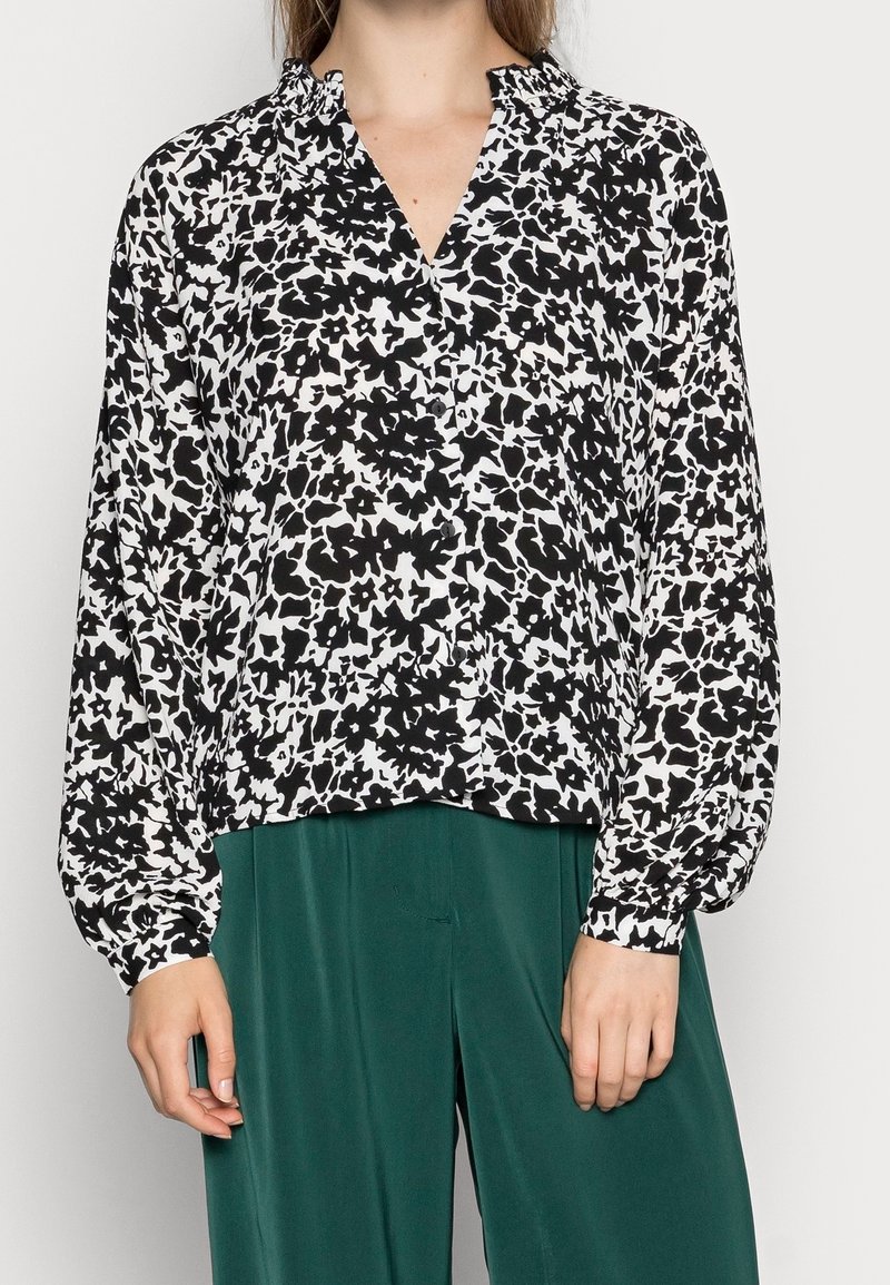 Blouse à imprimé floral noir et blanc avec un col en V, des manches longues et un décolleté froncé, associée à un pantalon vert. Tissu doux et léger.