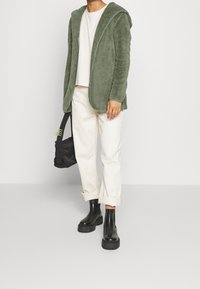 Green fleece hoodie, white t-shirt, rolled white pants, and black platform ankle boots. Holds a black bag with green accents.