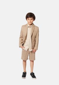 Completo giacca beige e pantaloni corti in tessuto testurizzato, abbinato a una camicia button-up beige chiara e sneakers nere.