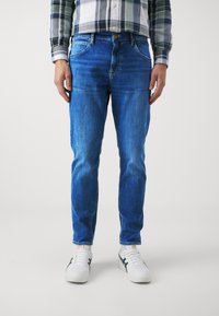 Pantalones vaqueros de denim azul con un diseño ajustado, que presentan un ligero desgaste, estilo de cinco bolsillos y cierre de botón estándar. Lleva puestos unos zapatillas blancas.