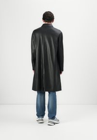 Schwarze Lederjacke mit geradem Schnitt, langen Ärmeln und einem Schlitzausschnitt hinten; kombiniert mit blauen Jeans und weißen Sneakers. Minimalistisches Design.