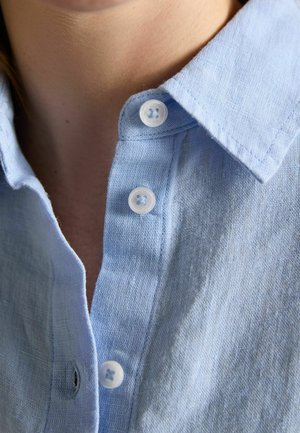 Kołnierzyk jasnoniebieskiej lnianej koszuli z trzema przezroczystymi guzikami, częściowo odpięty, na tle nagiej skóry szyi.