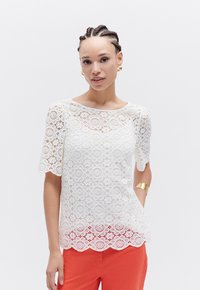 Top in pizzo bianco con maniche corte, motivo floreale, orlo a onde e scollatura rotonda. Abbinato a pantaloni arancioni.