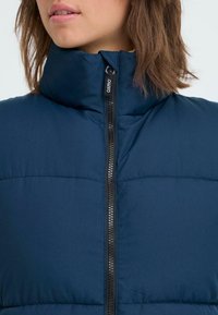 Tums zilas puffer veste ar augstu apkakli, kas ir aprīkota ar priekšējo rāvējslēdzēju un horizontāliem šuvēm. Materiāls izskatās mīksts un piepildīts.
