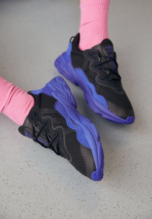 Osoba nosící černo-fialové sportovní tenisky s tlustými podrážkami a růžové žebrované ponožky na šedé mramorované podlaze.