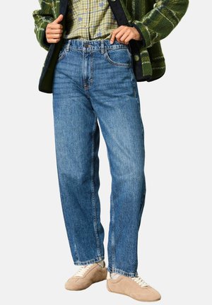 Person trägt blaue Jeans mit geradem Bein, beige Sneaker, ein grünes kariertes Hemd und eine grüne Fleecejacke vor einem einfarbigen Hintergrund.