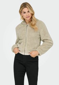 Light beige faux fur zip-up jacket with a hood, ribbed hem, and cuffs; features soft texture and a relaxed fit. Model wears black jeans.