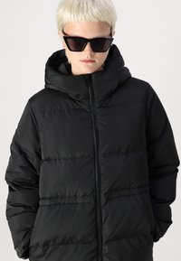 Juoda pūkine striukė su kapuce, su užtrauktuku ir pernapuku. Pagaminta iš lygaus, lengvo audinio.