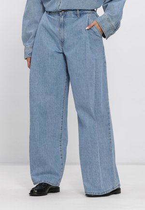 Hellblaue Jeans mit hoher Taille und weitem Bein, getragen mit schwarzen Schuhen und einem passenden hellblauen Jeanshemd, eine Hand in der Tasche.