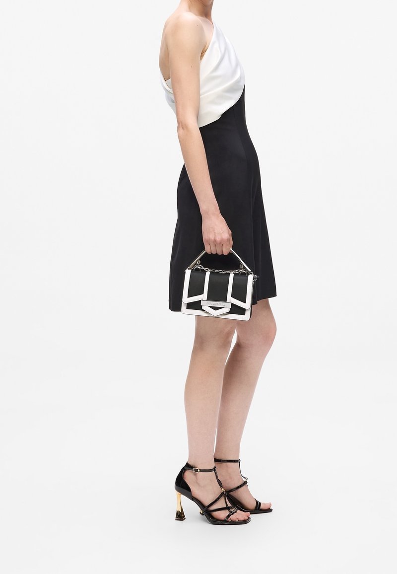 Femme en robe noire et blanche sans manches tenant un sac à main noir et blanc, portant des sandales noires à talons aiguille avec des talons dorés.
