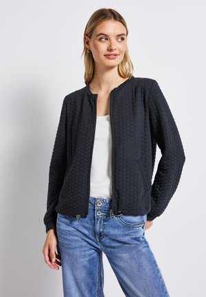 Jeune femme portant une veste matelassée noire zippée, un haut blanc et un jean bleu, se tenant avec une main dans la poche, regardant sur le côté.