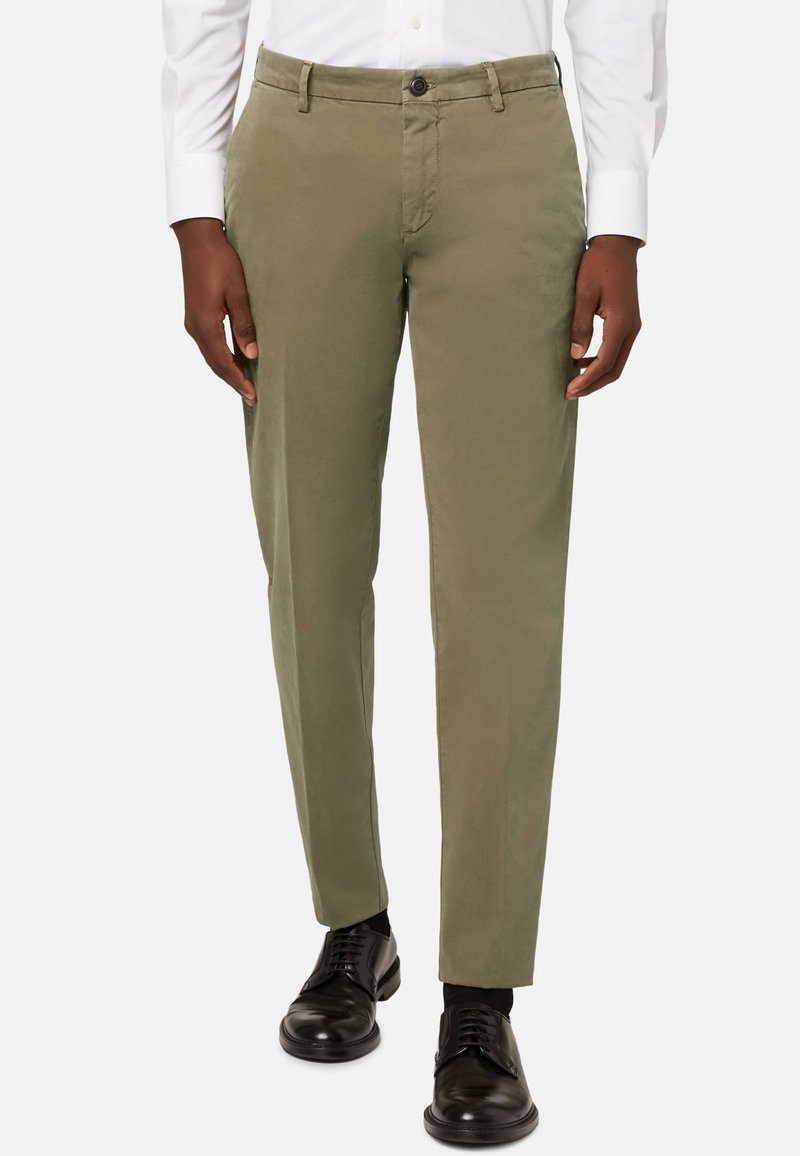 Olivgrüne Chinos aus Baumwollmischgewebe im klassischen Schnitt, mit seitlichen Taschen und einem Knopfverschluss. Getragen mit schwarzen Schnürschuhen.