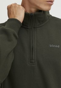 Sudadera de forro polar verde oliva con cuello de media cremallera, mangas acanaladas y detalles cosidos. Presenta un sutil logotipo impreso en el pecho.