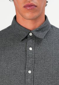 Grå skjorte med knepping i strukturet stoff, krage og fem hvite knapper foran. Nærbilde av øvre torso.
