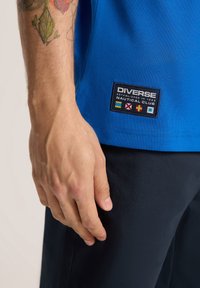 Niebieska bawełniana koszula z naszywką przedstawiającą "Diverse Nautical Club" oraz flagi. Posiada krótkie rękawy i gładką teksturę.