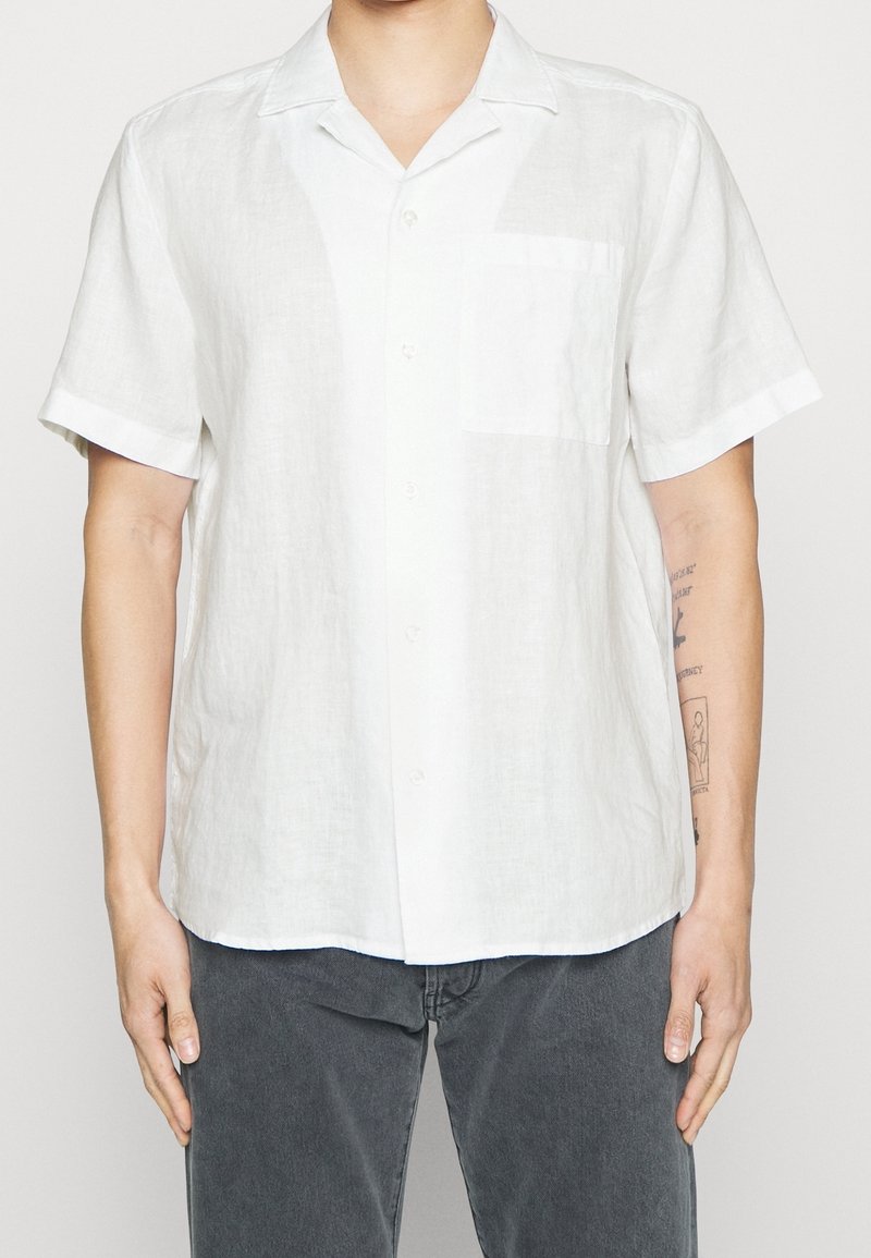 Homme portant une chemise blanche à manches courtes, boutonnée avec une poche poitrine unique, et un pantalon gris foncé, debout avec les bras détendus.