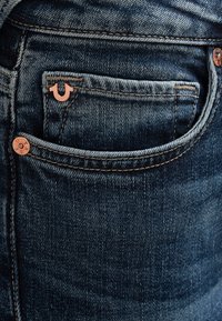 Jean en denim foncé avec un gros plan sur la poche arrière. Les caractéristiques incluent des coutures claires, des accents en métal, et une petite étiquette logo.