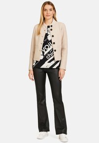 Beżowa krótka kurtka z okrągłym kołnierzem i czarnymi guzikami, noszona na czarno-białej koszulce z grafiką. W komplecie z czarnymi spodniami i białymi trampkami.