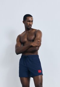 Tmavě modré plavecké šortky s elastickým pasem, šňůrkou na stažení a červenou značkovou nášivkou. Materiál působí lehce a hladce. Model je bez trička.