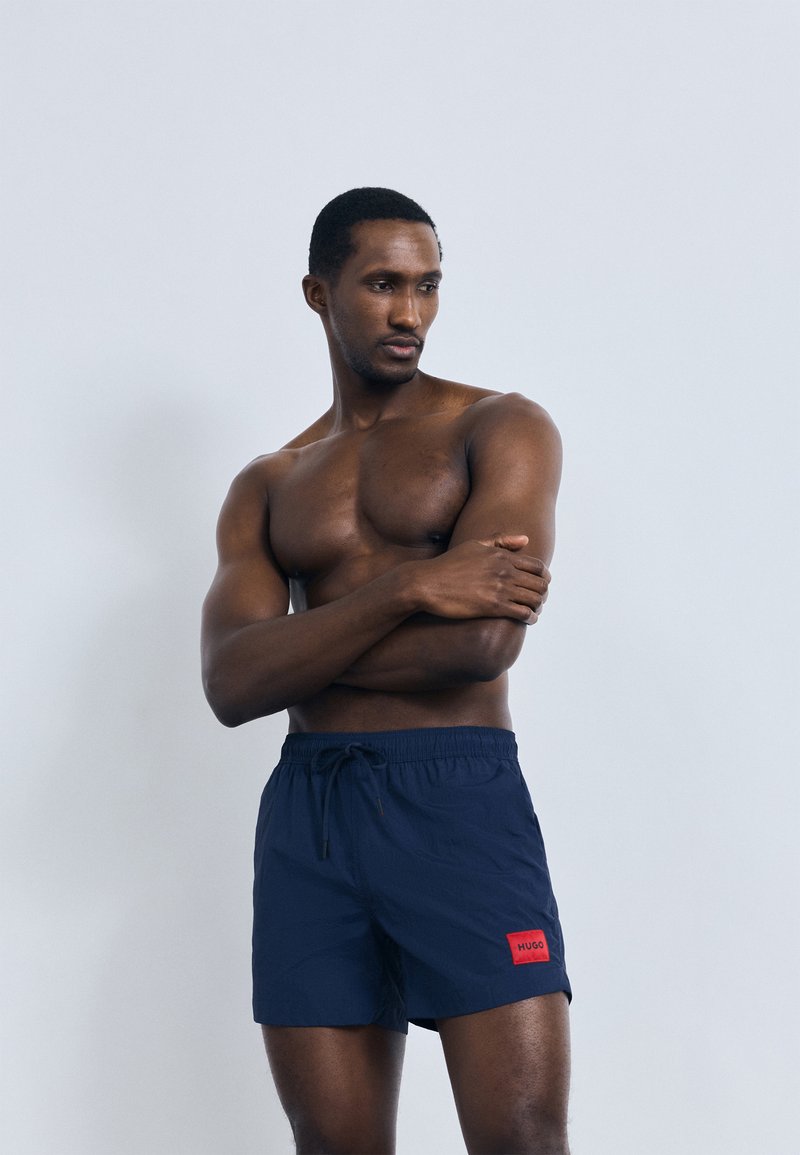 Tmavě modré plavecké šortky s elastickým pasem, šňůrkou na stažení a červenou značkovou nášivkou. Materiál působí lehce a hladce. Model je bez trička.