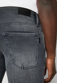 Šedé džíny z denimu s kapsou na zadní straně zdobenou prošíváním a černou štítkovou páskou na pasu. Textura je hladká s mírným vyblednutím.