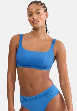 Woman with braided hair wearing a blue two-piece swimsuit with decorative beads on straps and waistband against a plain background.