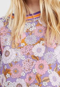 Blonde persoon draagt een paarse top met een gedetailleerd bloemenpatroon in tinten van roze, wit en oranje.