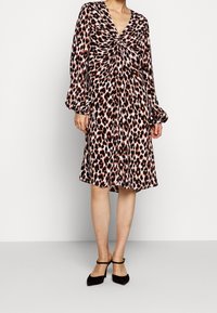 Kobieta w sukience w leopardzi wzór do kolan, z długimi rękawami i skręconym detalem z przodu, w czarnych szpilkach na cienkich paskach.