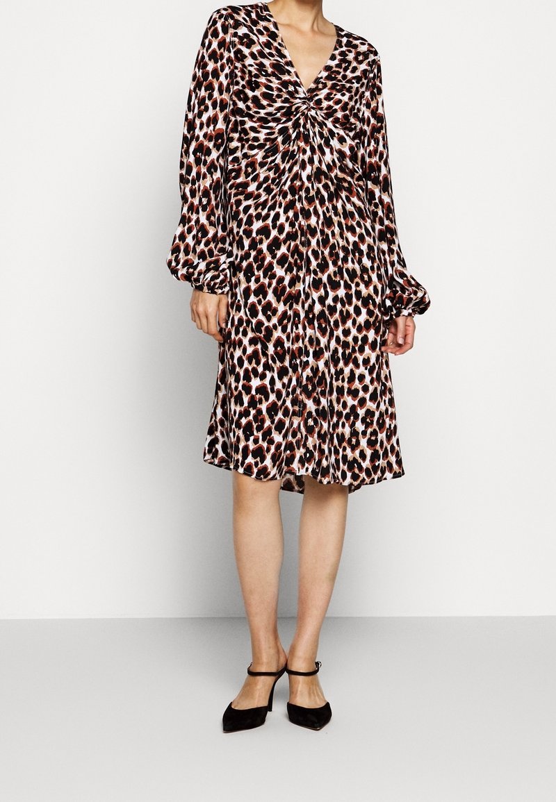 Kobieta w sukience w leopardzi wzór do kolan, z długimi rękawami i skręconym detalem z przodu, w czarnych szpilkach na cienkich paskach.
