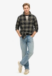 Geruite flanellen shirt in donkergroen en marineblauw, gedragen over een wit T-shirt, gecombineerd met lichtblauwe jeans en beige sneakers. Casual pasvorm.