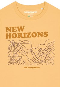 Gele katoenen t-shirt met een geprinte illustratie van bergen, een wandelaar, en de tekst "NIEUWE HORIZONTEN ...zijn overal" in bruin.