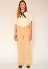 Chemise beige oversize à manches courtes, associée à un pantalon flared en velours côtelé jaune clair taille haute. Les accessoires comprennent une écharpe noire et des bracelets.