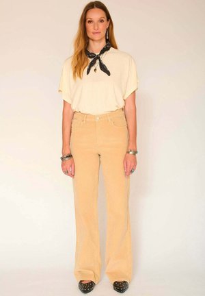 Chemise beige oversize à manches courtes, associée à un pantalon flared en velours côtelé jaune clair taille haute. Les accessoires comprennent une écharpe noire et des bracelets.