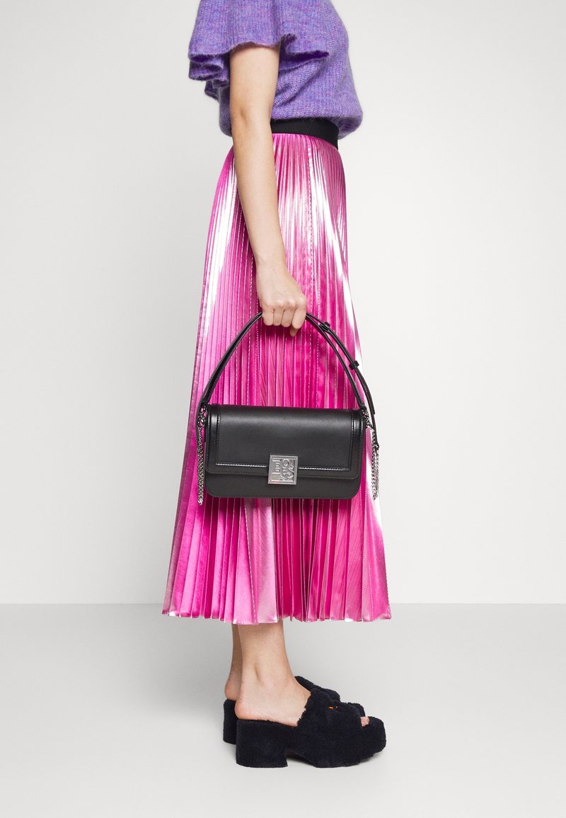 Femme portant un pull violet et une jupe plissée rose, tenant un sac à main noir, chaussée de mules à talons noires et poilues à bout ouvert, debout de côté.