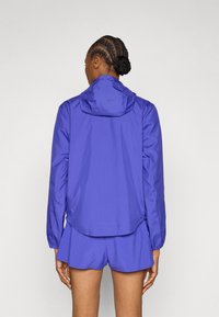 Μωβ ελαφρύ τζάκετ με κουκούλα, με λεία υφή, μακριά μανίκια και ελαφρώς καμπυλωτό τελείωμα. Συνδυασμένο με σορτς στο ίδιο χρώμα.