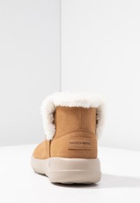 Stivale alla caviglia in suede marrone con colletto in pelliccia sintetica bianca, suola flessibile di un beige chiaro e logo Skechers sul lato.