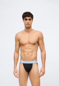 Jonge man met kort donker haar, gekleed in zwarte onderbroek met een lichtblauwe Calvin Klein tailleband, staande tegen een egaal witte achtergrond.