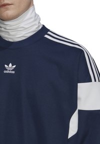 Mörkblå sweatshirt med vita accenter och tre vita ränder på axlarna. Har en rund hals och Adidas-logotyp på bröstet.
