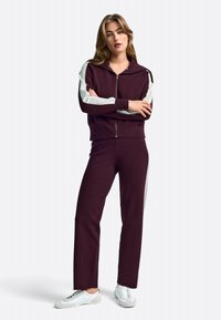 Bordo športni trenirka z jopico na zadrgo, okrašena s svetlo sivimi poudarki, in ujemajoče se hlače. Obuta je z belimi supergami, tkanina je gladka na otip.