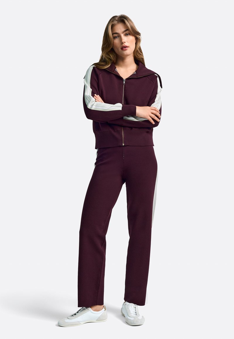 Bordo športni trenirka z jopico na zadrgo, okrašena s svetlo sivimi poudarki, in ujemajoče se hlače. Obuta je z belimi supergami, tkanina je gladka na otip.