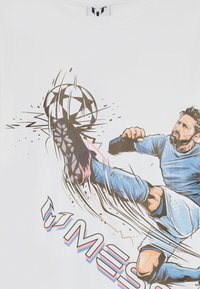 Hvit t-skjorte med et fargerikt grafisk motiv av en spiller som sparker en fotball, med dynamiske linjer og teksten "Messi" i fete, stiliserte bokstaver.