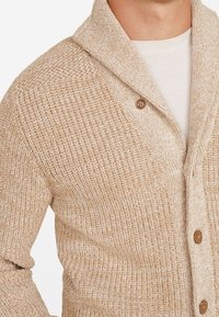 Cardigan beige en tricot avec un motif texturé, col châle, fermeture à un bouton, et tissu doux. Présente une coupe décontractée.