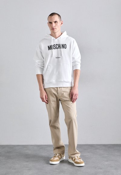 Hanorac alb cu glugă, cu logo-ul negru "Moschino Milano", asortat cu pantaloni bej și sneakers maro și alb. Material neted și croială lejeră.