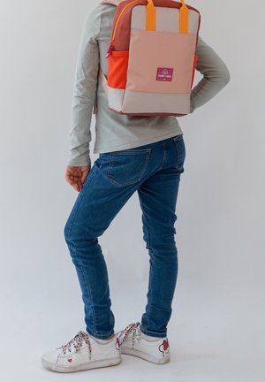 Personne portant un jean bleu et des baskets blanches avec des détails en forme de cœur, portant un sac à dos rose et orange avec des bretelles jaunes.
