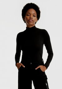 Zwarte turtleneck met lange mouwen en een aansluitend ontwerp, gemaakt van gladde stof. Het model draagt zwarte broek met zichtbare zakken en ritsen.