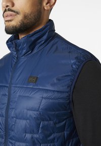 Chaleco aislante azul marino con cuello alto, diseño acolchado y parche de logo negro en el pecho. Textura suave y cierre con cremallera.